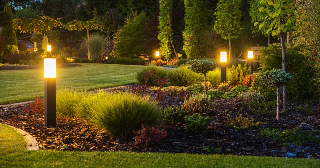 Jardin paysager illuminé la nuit par des bornes lumineuses modernes. Pelouse verte impeccable et parterres de plantes variées, créant une ambiance chaleureuse.