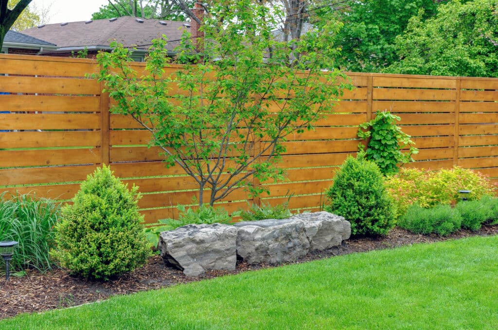 Clôture en bois horizontale délimitant un jardin paysager avec un jeune arbre, des arbustes et de grandes roches décoratives devant une pelouse verte.