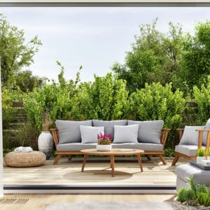 Patio moderne avec salon de jardin en bois: canapé, fauteuil, table basse. Coussins gris, pouf osier, verdure dense.