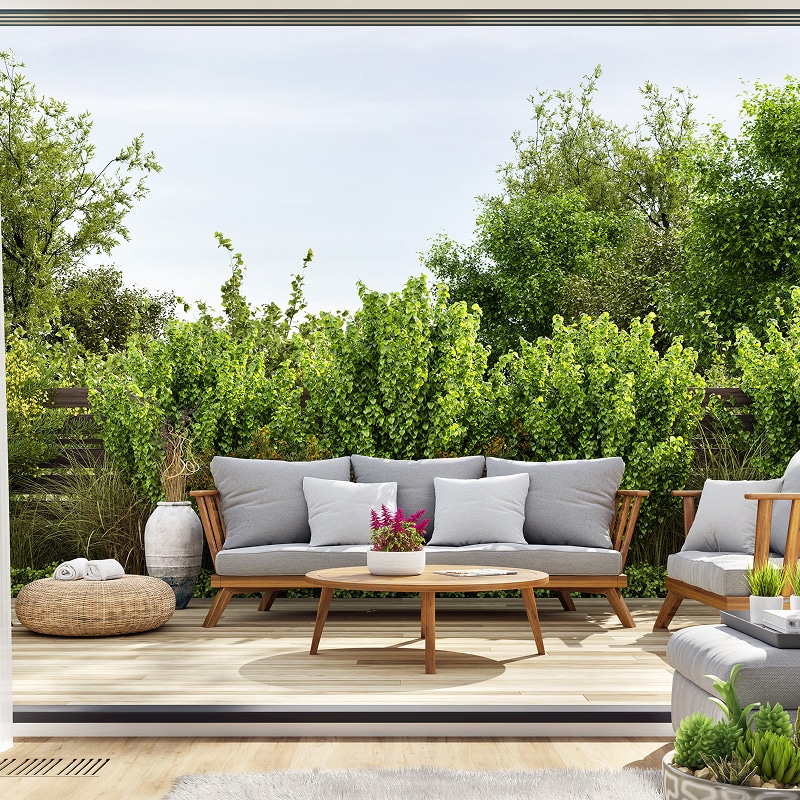 Patio moderne avec salon de jardin en bois: canapé, fauteuil, table basse. Coussins gris, pouf osier, verdure dense.