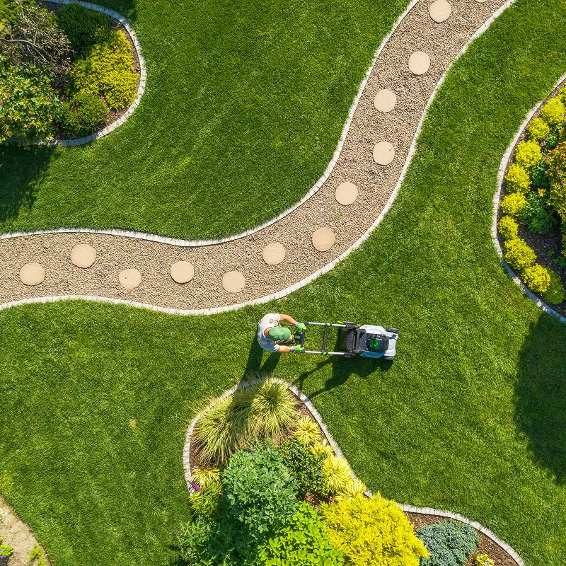 Tonter la pelouse: Entretien professionnel du jardin Vue aérienne: homme tondant pelouse luxuriante. Allée sinueuse en gravier et pas japonais traverse jardin avec massifs fleuris.